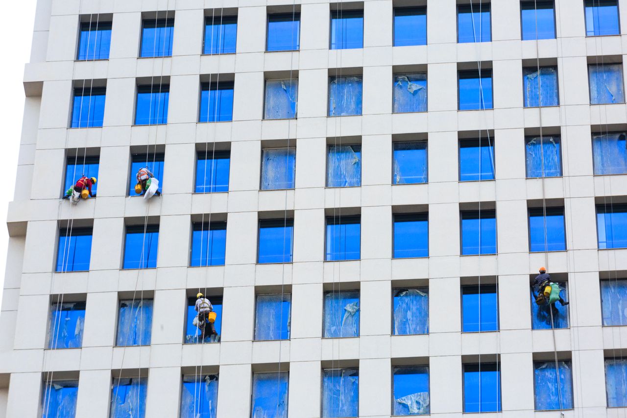 services-01 Des professionnels nettoient les vitres d’un gratte-ciel à l’aide de cordes, en milieu urbain.